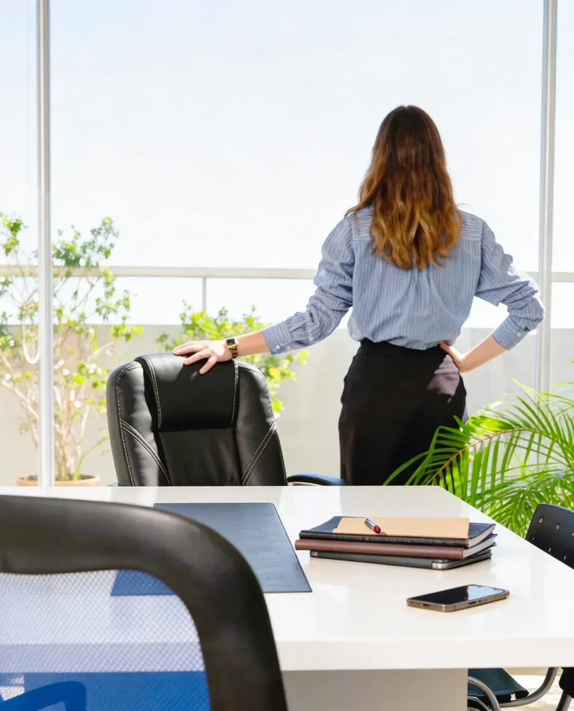 Eine Geschäftsfrau blickt aus einem hellen Bürofenster auf eine Terrasse; symbolisiert Weitblick und Kontrolle über das Unternehmen.