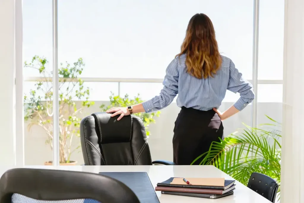 Eine Geschäftsfrau blickt aus einem hellen Bürofenster auf eine Terrasse; symbolisiert Weitblick und Kontrolle über das Unternehmen.